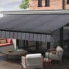 Retractable striped fabric awning, dark grey with diagonal pattern and striped valance, fully extended over sliding glass doors—shading a sunlit modern patio with wooden deck, cushioned seating and coffee table.
